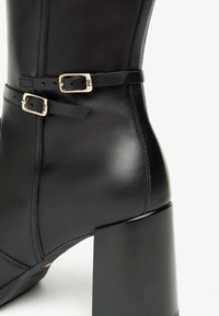 Schwarze LederStiefelette mit einem quadratischen Absatz, ausgestattet mit zwei verstellbaren Riemen mit goldenen Schnallen und detaillierter Naht auf der Seite.