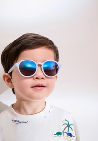 Lunettes de soleil rondes bleu clair avec des verres miroirs. L'enfant porte un sweat-shirt blanc avec une broderie colorée de dinosaures et un motif de palmier.