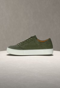 Πράσινο σουέτ αθλητικό παπούτσι με λευκή καουτσούκ σόλα, στρογγυλεμένη μύτη, έξι οπές για κορδόνια και καφέ εσωτερική επένδυση. Απλός σχεδιασμός με ελάχιστες λεπτομέρειες.