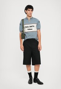 Polo azul de manga corta con una franja blanca que presenta el texto "PAUL SMITH" y "Cicilismo". Llevado con pantalones cortos negros y zapatos negros, se complementa con una bolsa verde.