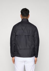 Melnā vieglā jaka ar augstu apkakli, sieta paneli un rāvējslēdzēja kabatu mugurā. Gluda tekstūra un vienkāršs dizains.