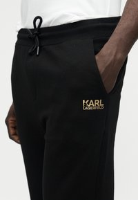 Zbliżenie osoby noszącej czarne spodnie dresowe Karl Lagerfeld ze złotym logo przy kieszeni, ręką w kieszeni oraz białą koszulą włożoną do środka.