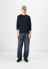 Pull marin, casquette gris clair, jean en denim oversized en teinte foncée, et chaussures noires. Design simple, texture douce et coupe décontractée.