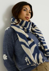 Bufanda a rayas en azul marino y crema hecha de tela suave, con bordes con flecos, colocada alrededor del cuello de una persona que lleva un suéter azul texturizado.