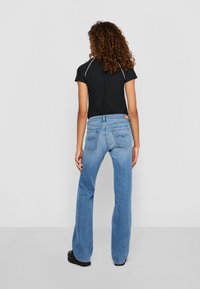 Kvinne med krøllete hår står med ryggen til iført en svart t-skjorte med korte ermer, lyseblå bootcut jeans og svarte sko på en ensfarget bakgrunn.