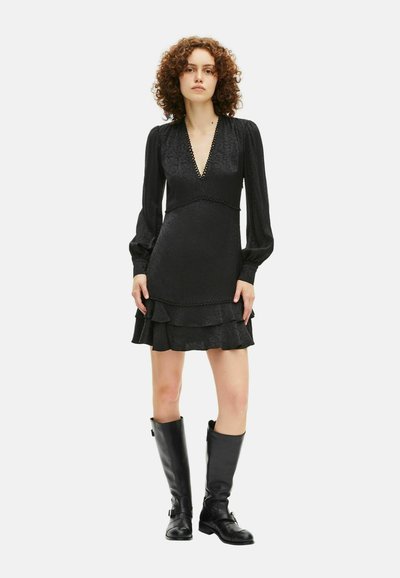 Kvinne med krøllete hår iført en svart langermet V-hals mini kjole med volanger og svarte skinnstøvler til knærne, står på hvit bakgrunn.