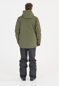 Wasserdichte grüne Jacke mit Kapuze, mit lockerer Passform und verstellbaren Manschetten, kombiniert mit lockeren schwarzen Schneehosen und Schneestiefeln.