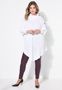 Witte gebreide poncho met een colkraag, met textuurpatronen en zijknopen, gecombineerd met strakke donkerpaarse leggings en zwarte hakken.