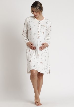 Mujer embarazada que lleva un camisón premamá blanco de manga larga con estampados de animales, de pie descalza, con las manos suavemente sobre el vientre, sobre un fondo liso.