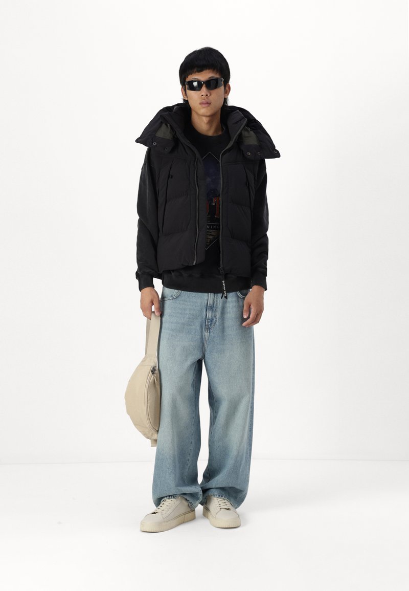 Chaleco acolchado negro sobre un sudadero negro, jeans de pierna ancha azul claro, zapatillas blancas y un bolso de hombro beige. Los accesorios incluyen gafas de sol.