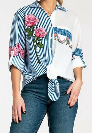 Vrouw draagt een blauw-wit gestreept overhemd met knopen met roze rozenborduursels en een blauwe spijkerbroek, overhemd in de taille gestrikt, mouwen opgerold.
