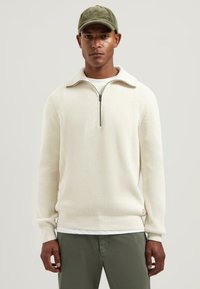 Geribbeld wit sweatshirt met een ritskraag, gedragen over een wit shirt. Combineer met olijfgroen broek, met een klein logo op de zoom.