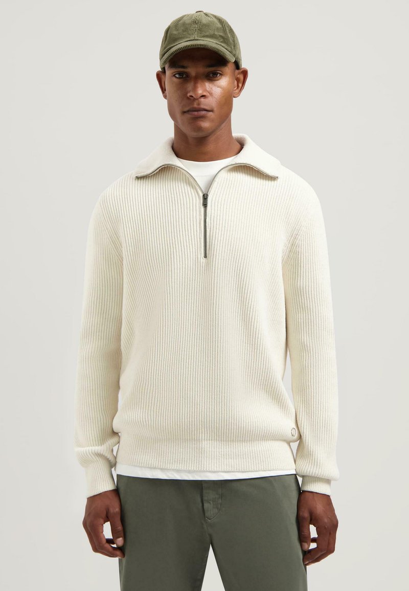 Geribbeld wit sweatshirt met een ritskraag, gedragen over een wit shirt. Combineer met olijfgroen broek, met een klein logo op de zoom.