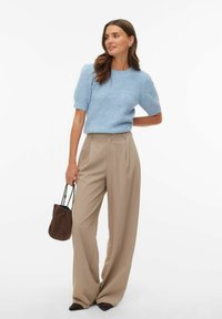Femme portant un pull en tricot à manches courtes bleu clair, un pantalon beige à jambes larges, des chaussures pointues marron et tenant un petit sac à main marron.