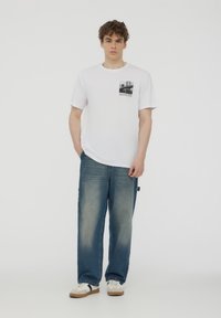 T-shirt bianco a maniche corte con una grafica in bianco e nero sul lato sinistro del petto. Abbinato a jeans azzurri chiari, a taglio morbido, e sneakers bianche.