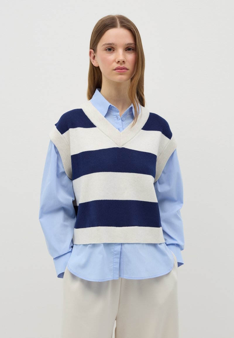 Gilet a righe navy e crema senza maniche con scollatura a V, indossato sopra una camicia azzurra a bottoni, abbinato a pantaloni crema.