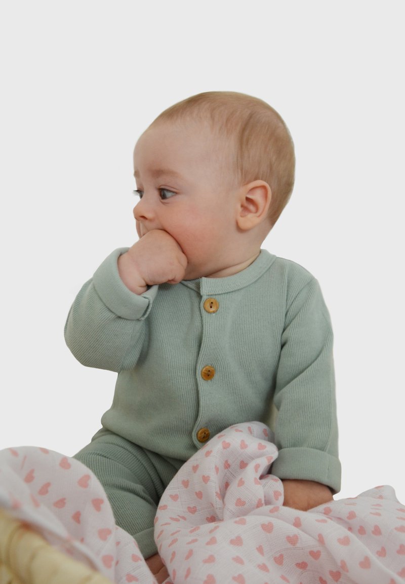 Grüner, gestreifter Baby-Overall mit Holzknöpfen. Sitzt auf einer hellrosa Decke mit Herzmuster und hält eine Hand zum Mund.