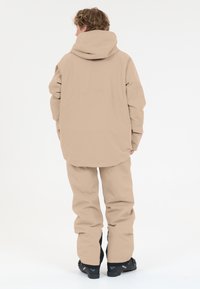 Beige vedenpitävä takki ja housut, joissa on rento malli, huppu ja vahvistetut hihansuut. Sileä rakenne hienovaraisilla saumoilla ja ompeluilla.