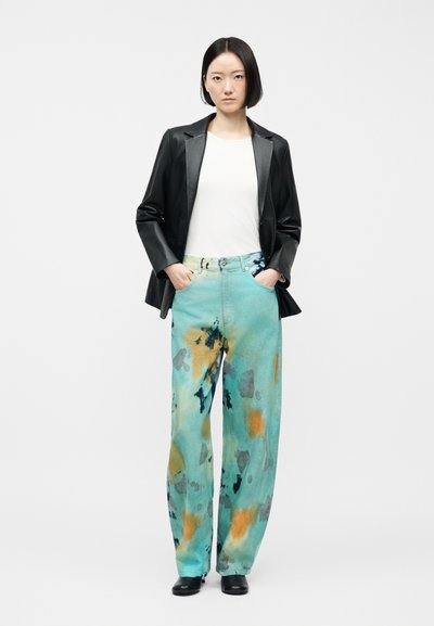 Γυναίκα με μαύρο καρέ χτένισμα, φορώντας μαύρο δερμάτινο σακάκι, λευκή μπλούζα, φαρδιά πλεκτά μπλε παντελόνια με μοτίβο tie-dye, μαύρα παπούτσια, στέκεται με τα χέρια στις τσέπες.