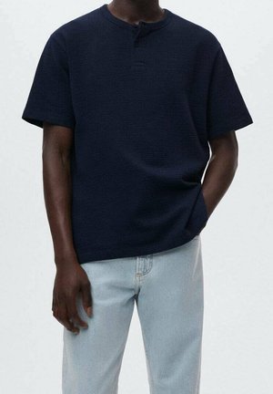 Granatowy krótki t-shirt z teksturowanej tkaniny, z okrągłym dekoltem i pojedynczym guzikiem przy kołnierzu, zestawiony z jasnymi dżinsami.