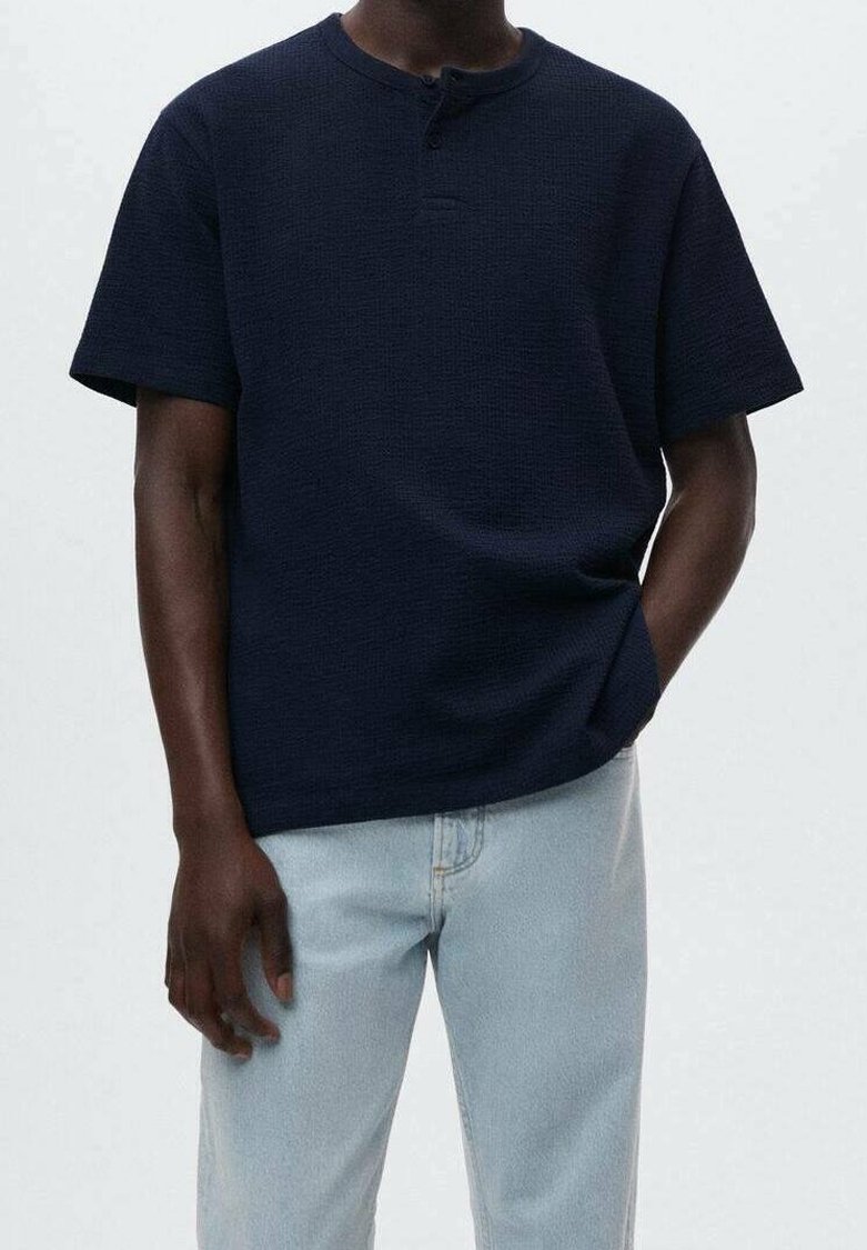 T-shirt bleu marine à manches courtes avec un tissu texturé, présentant un col rond et un bouton unique au col, associé à un jean clair.