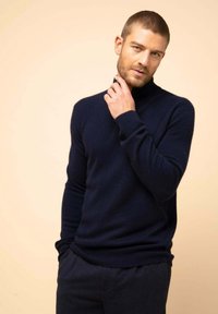 Granatowy sweter z golfem wykonany z miękkiego materiału dzianinowego, z prążkowanymi mankietami i dołem, w połączeniu z ciemnymi spodniami na neutralnym tle.