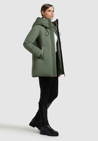 Giacca puffer verde con colletto alto, cordini, due tasche laterali e vestibilità comoda, abbinata a leggings neri e stivali al ginocchio.