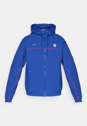Veste coupe-vent Nike bleue avec capuche, dotée d'accents rouges et d'un logo. La veste a une fermeture éclair et des poignets et un ourlet élastiques.