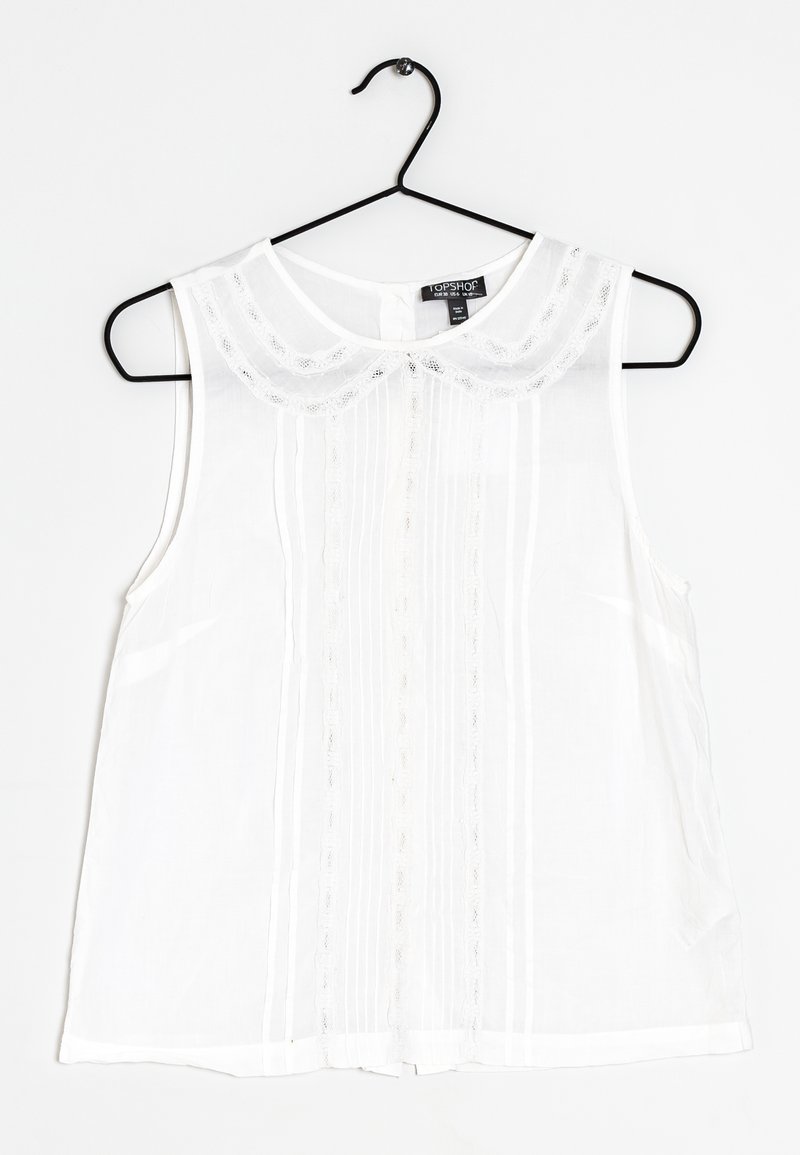 Blusa bianca senza maniche con colletto rotondo, bordi di pizzo e dettagli a righe verticali. Realizzata in tessuto leggero, appesa a un gancio nero.
