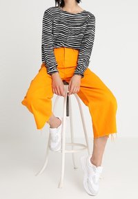 Černobíle pruhovaná dlouhá rukávová halenka v kombinaci s oranžovými širokými kalhotami s vysokým pasem a bílými teniskami se šedými akcenty.