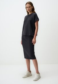 Czarna, oversize koszulka z podwiniętymi rękawami, zestawiona z dopasowaną spódnicą do kolan. Modelka ma na sobie białe trampki na jednolitym tle.