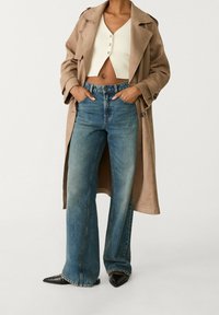 Kvinna i beige trenchcoat, krämfärgad kortärmad knäppt topp, vida blå jeans och svarta spetsiga skor med tårna pekande framåt, med händerna i fickorna.