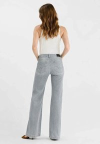 Femme aux longs cheveux bruns porte un haut blanc sans manches et un jean large gris clair, debout avec les mains dans les poches arrière.