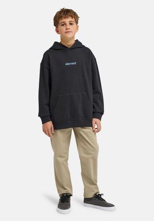 Garçon debout portant un sweat à capuche noir "element", un pantalon beige et des baskets noires à semelles blanches sur un fond blanc uni.