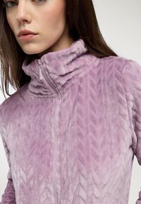 Giacca in pile viola con zip e colletto alto, caratterizzata da motivi a zigzag texture e una zip liscia. Materiale morbido e peloso.