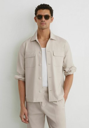 Mand iført beige skjorte med knapper over hvid tanktop, beige bukser og solbriller, stående med den ene hånd i lommen op ad en hvid væg.