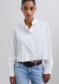 Camisa blanca de botones cortada con dobladillo y puños con volantes; hecha de una tela ligera; cuenta con un cuello clásico y una tapeta frontal.