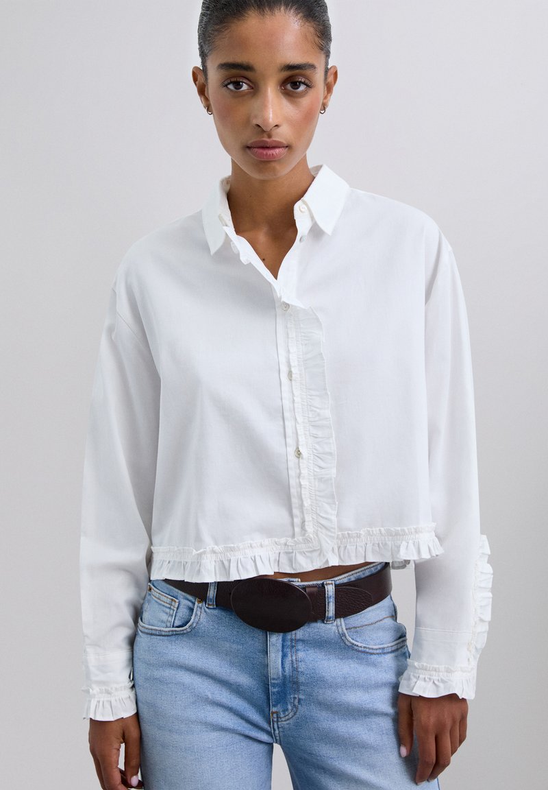 Camisa blanca de botones cortada con dobladillo y puños con volantes; hecha de una tela ligera; cuenta con un cuello clásico y una tapeta frontal.