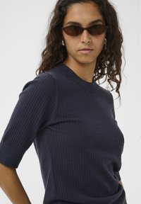 Marineblauer Strickpullover mit kurzen Ärmeln und Rundhalsausschnitt. Das Modell trägt eine dunkle Sonnenbrille und präsentiert eine glatte Textur sowie ein anliegendes Design.