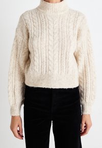 Cremefarbener Strickpullover mit hohem Kragen und gerippten Bündchen. Kurzer, oversized Schnitt mit strukturierten Mustern. Kombiniert mit schwarzen Cordhosen.