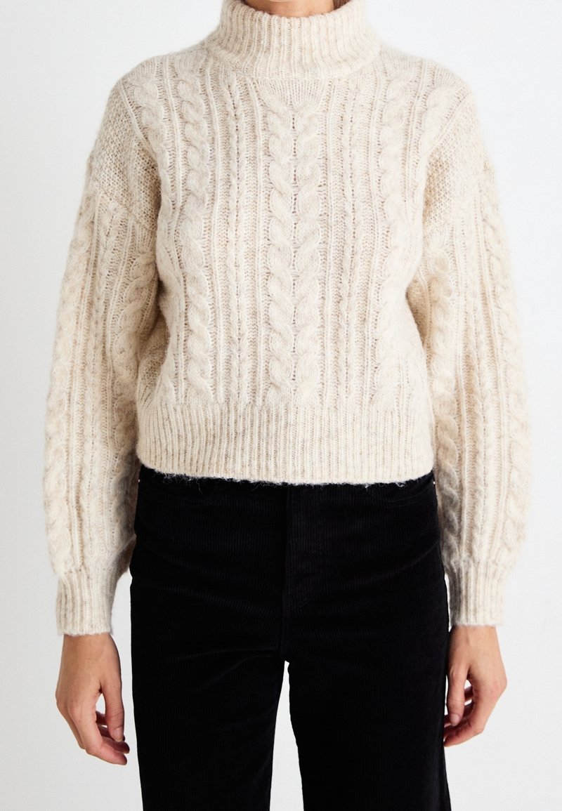 Cremefarbener Strickpullover mit hohem Kragen und gerippten Bündchen. Kurzer, oversized Schnitt mit strukturierten Mustern. Kombiniert mit schwarzen Cordhosen.
