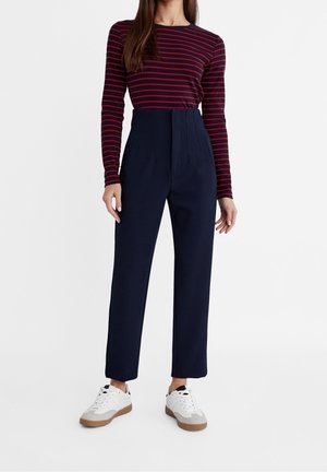 Vrouw draagt een rood-zwart gestreept top met lange mouwen, een marineblauwe broek met hoge taille en witte sneakers met bruine zolen.