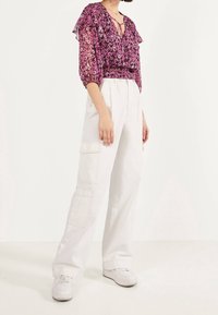 Kobieta w fioletowej bluzce w kwiaty z bufkami, białych spodniach cargo i białych trampkach, pozująca na jednolitym jasnym tle.