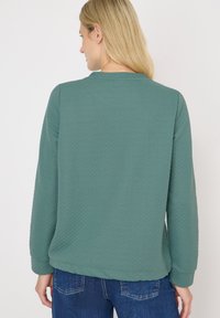 Kvinna med blont hår klädd i en texturerad grön långärmad topp och blå jeans, vänd bort mot en enkel vit bakgrund.
