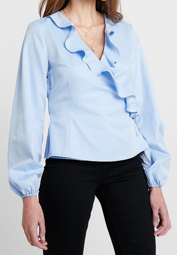 Femme portant un chemisier bleu clair à manches longues avec col en V volanté et pantalon noir, vue des épaules aux hanches.