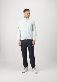 Casaco com fecho éclair de cor verde menta clara e padrão texturizado, combinado com calças azul-marinho. Ténis brancos completam o visual. Apresenta um logo no peito.