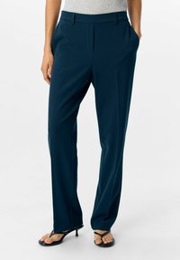 Femme portant un pantalon droit bleu marine, un haut sans manches gris, les mains dans les poches, et des sandales à talons noires sur un fond blanc.