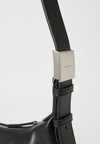 Čierna kožená crossbody taška s hladkým povrchom, vybavená kovovým zapínaním s gravírovaním "Calvin Klein" a nastaviteľným popruhom.