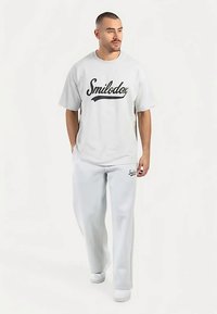 Camiseta blanca de gran tamaño con el logo negro "Smilodox", combinada con pantalones deportivos gris claro con un pequeño logo en el lado izquierdo. Zapatillas blancas.