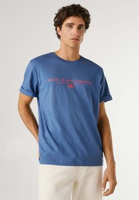 Ung mand med krøllet hår iført en blå "Pepe Jeans London" T-shirt og lyse bukser, stående foran en ensfarvet baggrund.